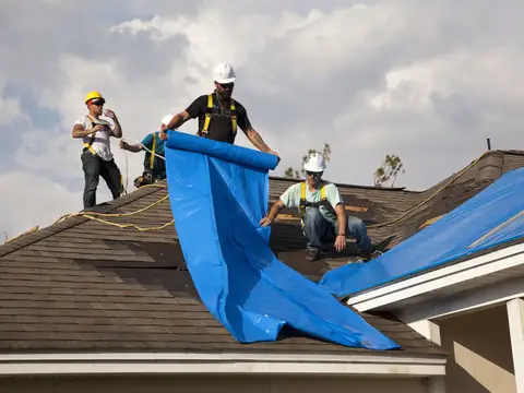 Workers in hard hats unrolling a blue tarp across a damaged house roof