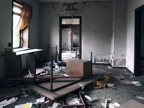 A wrecked room with broken furniture, fallen plaster and debris across the floor, daylight from a tall window